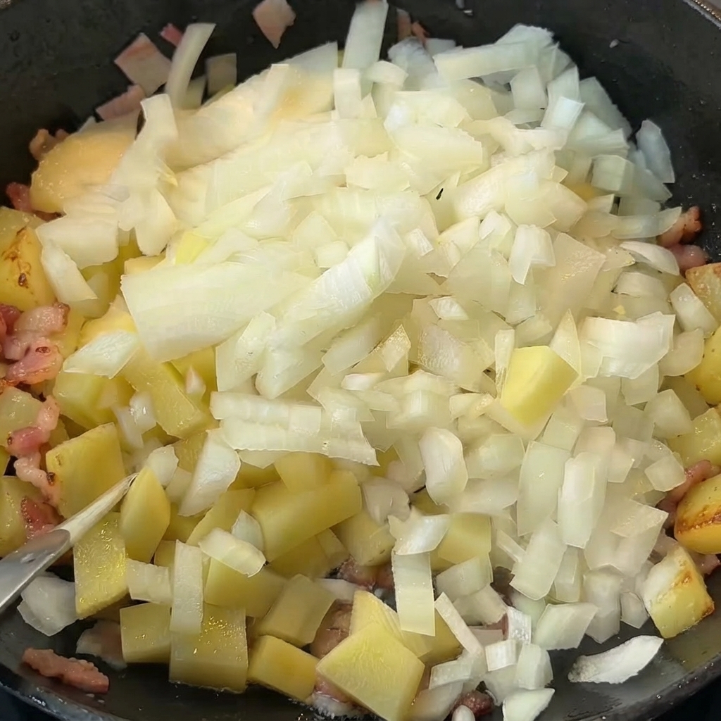 Cipolle a dadini versate sopra patate cotte e bacon in una padella.