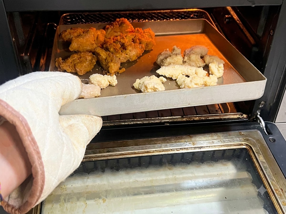 Un guanto da forno che tiene un vassoio pieno di popcorn chicken crudo o surgelato e ali piccanti allinterno di una friggitrice ad aria.