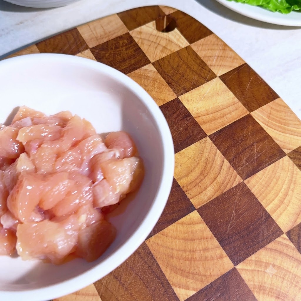Piccoli pezzi di pollo in una ciotola bianca con le spezie dosate cosparse sopra prima di essere mescolati.