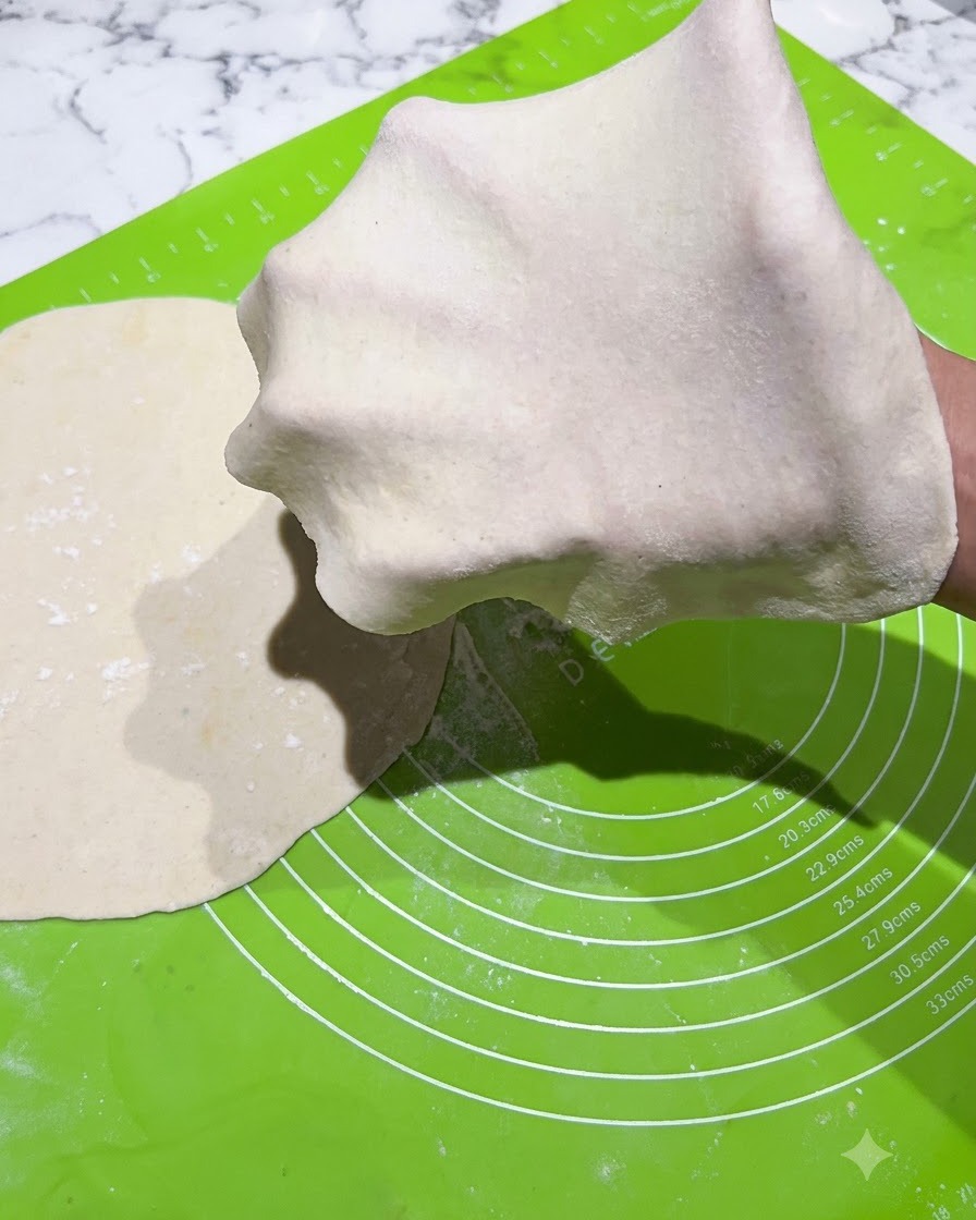Un sottile foglio di pasta stesa tirato sopra una mano guantata su un tappetino verde.