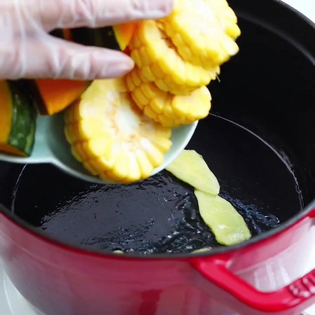 Pezzi di zucca e pannocchie di mais versati da un piatto nella pentola rossa.