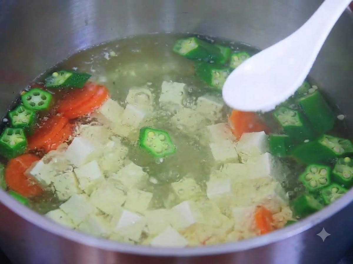 Un cucchiaio bianco che contiene sale sopra una pentola fumante di zuppa di tofu, okra e carote.