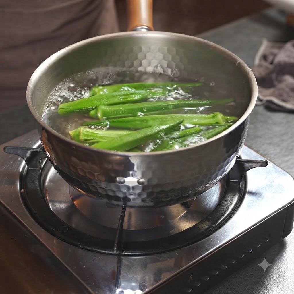 Baccelli di okra verde che bollono allinterno di una pentola metallica piena dacqua su un fornello.