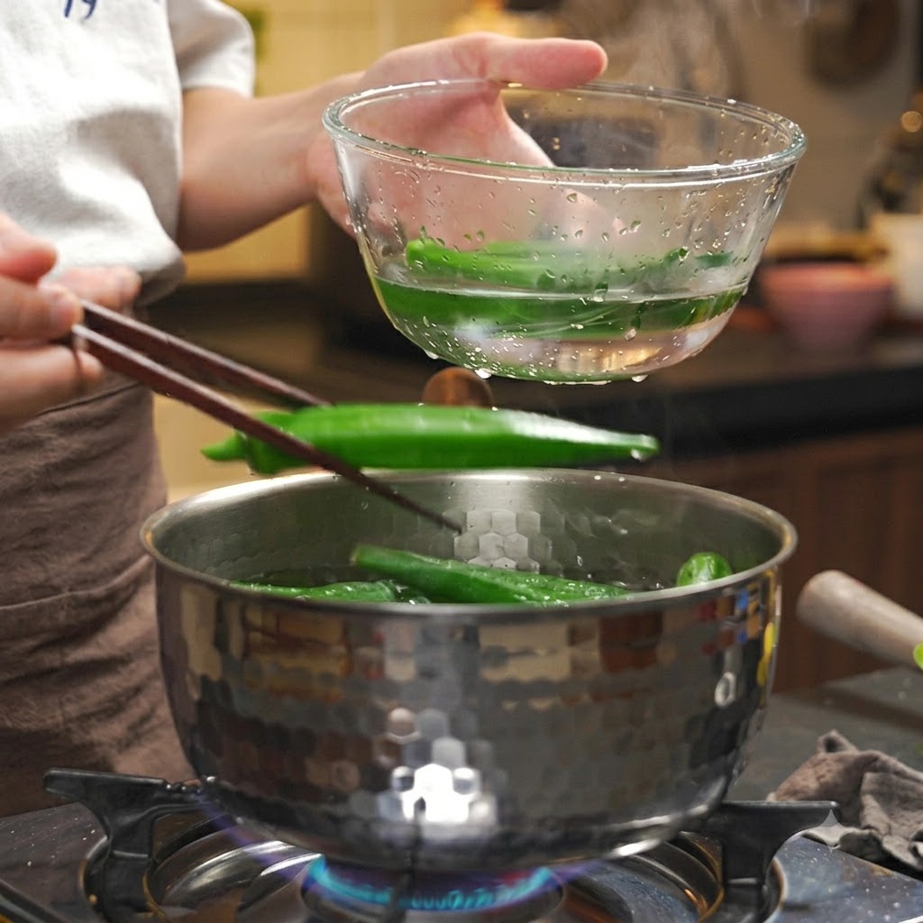 Okra cotta prelevata con le bacchette da una pentola calda in una ciotola trasparente di acqua ghiacciata.