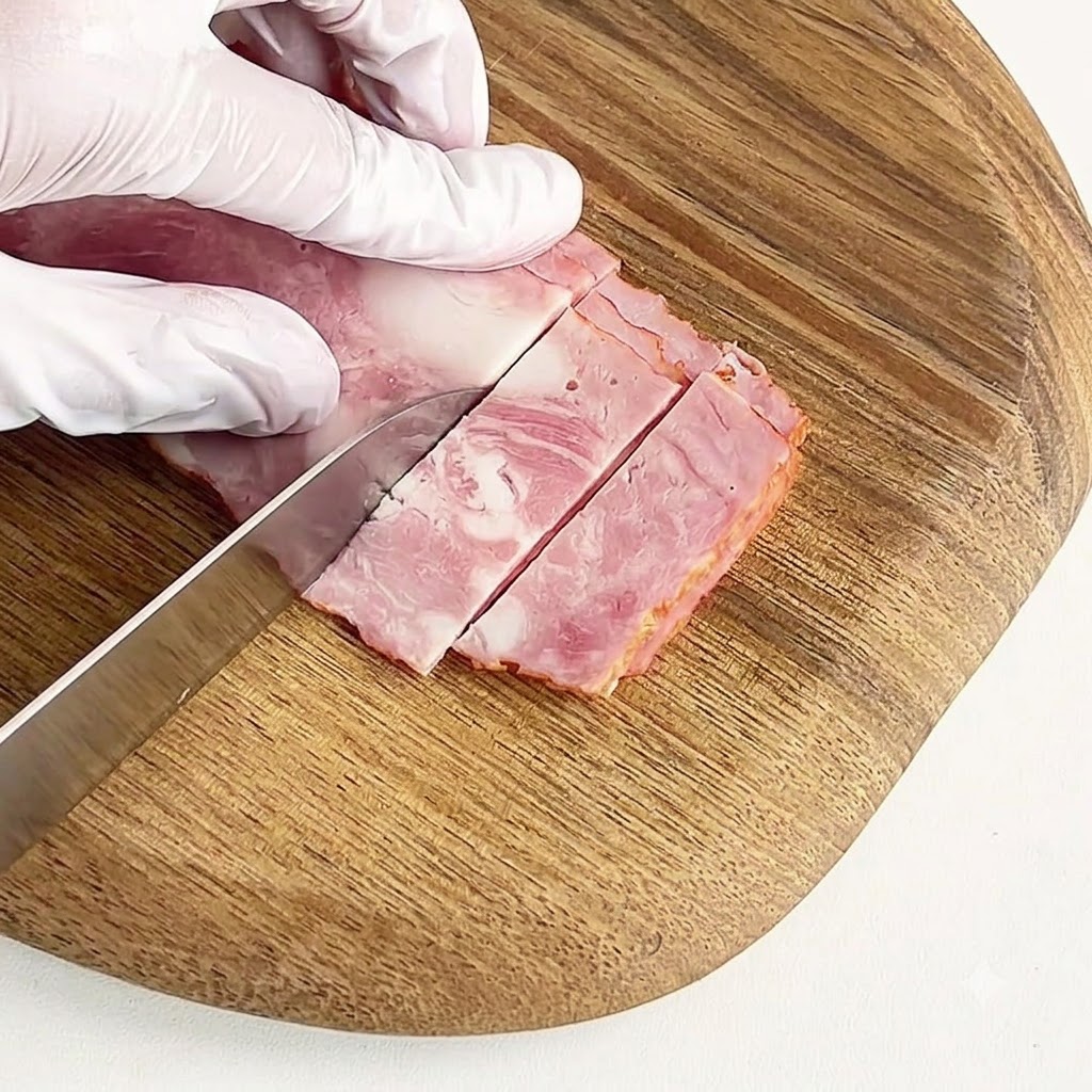 Una mano guantata che taglia la pancetta stagionata in piccoli pezzi su un tagliere di legno rotondo.