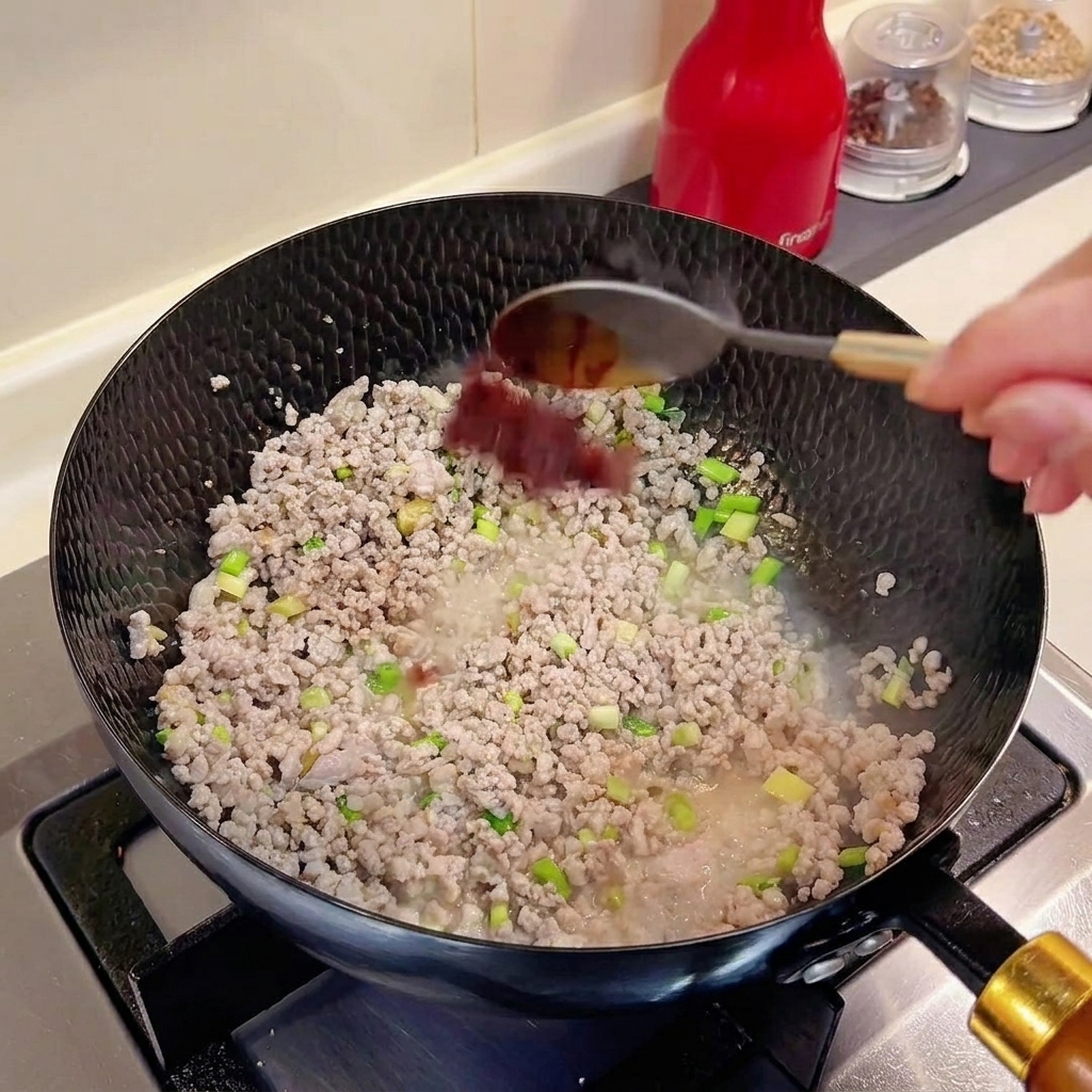 Un cucchiaio di doubanjiang aggiunto in un wok contenente carne macinata e aromi.