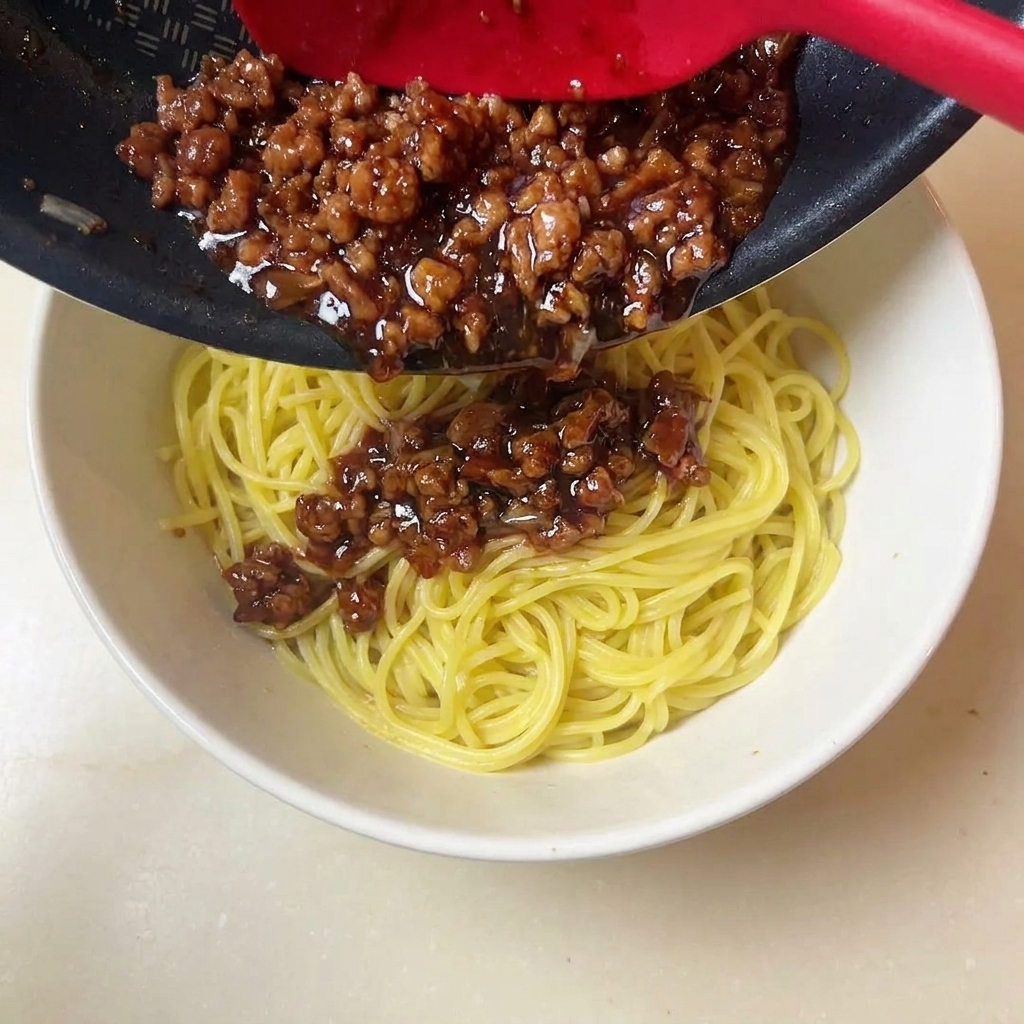 Una spatola rossa che raccoglie salsa di carne macinata ricca e scura su una ciotola di noodles gialli.