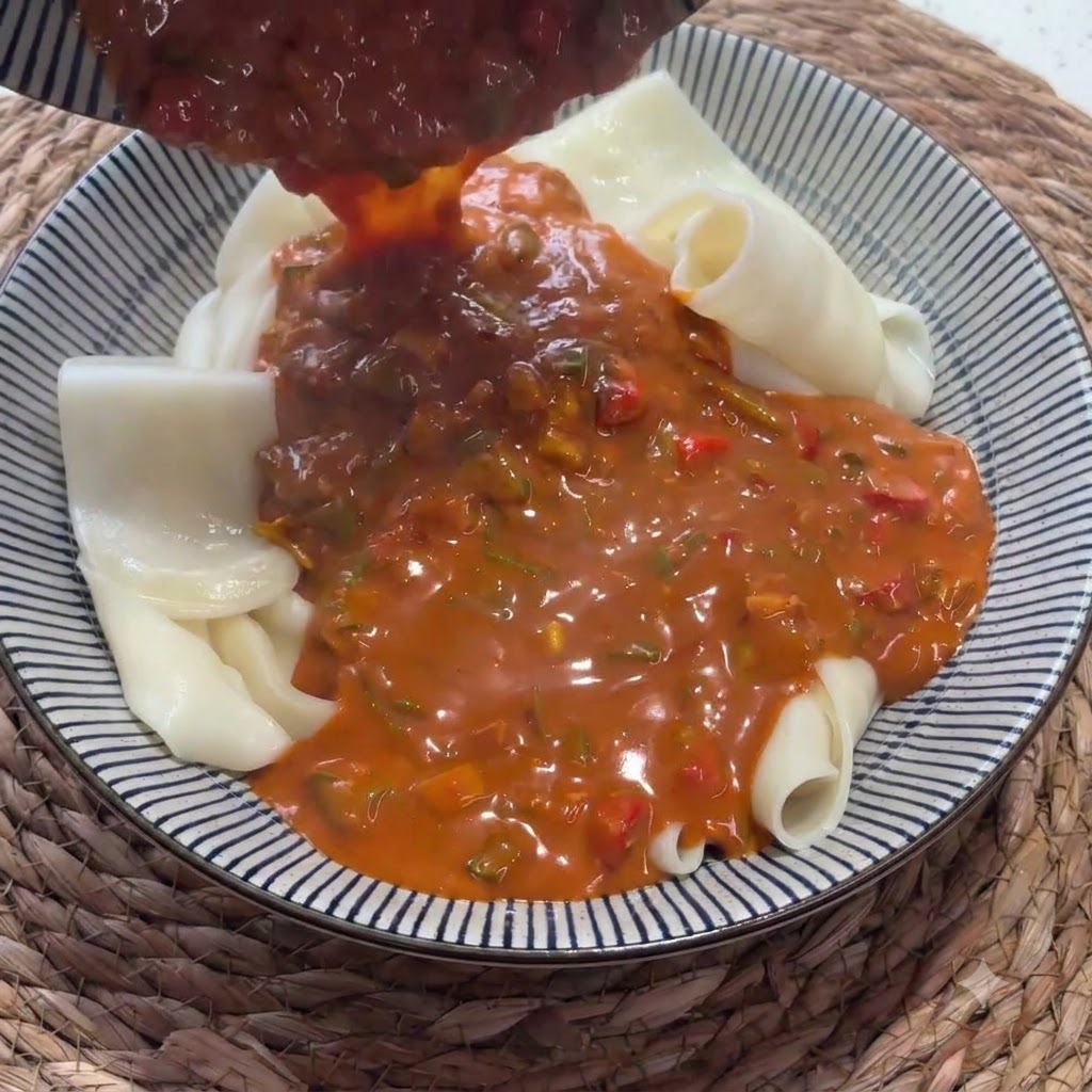 Salsa rosso vivo versata da un mettolino su una ciotola piena di tagliolini larghi piegati.
