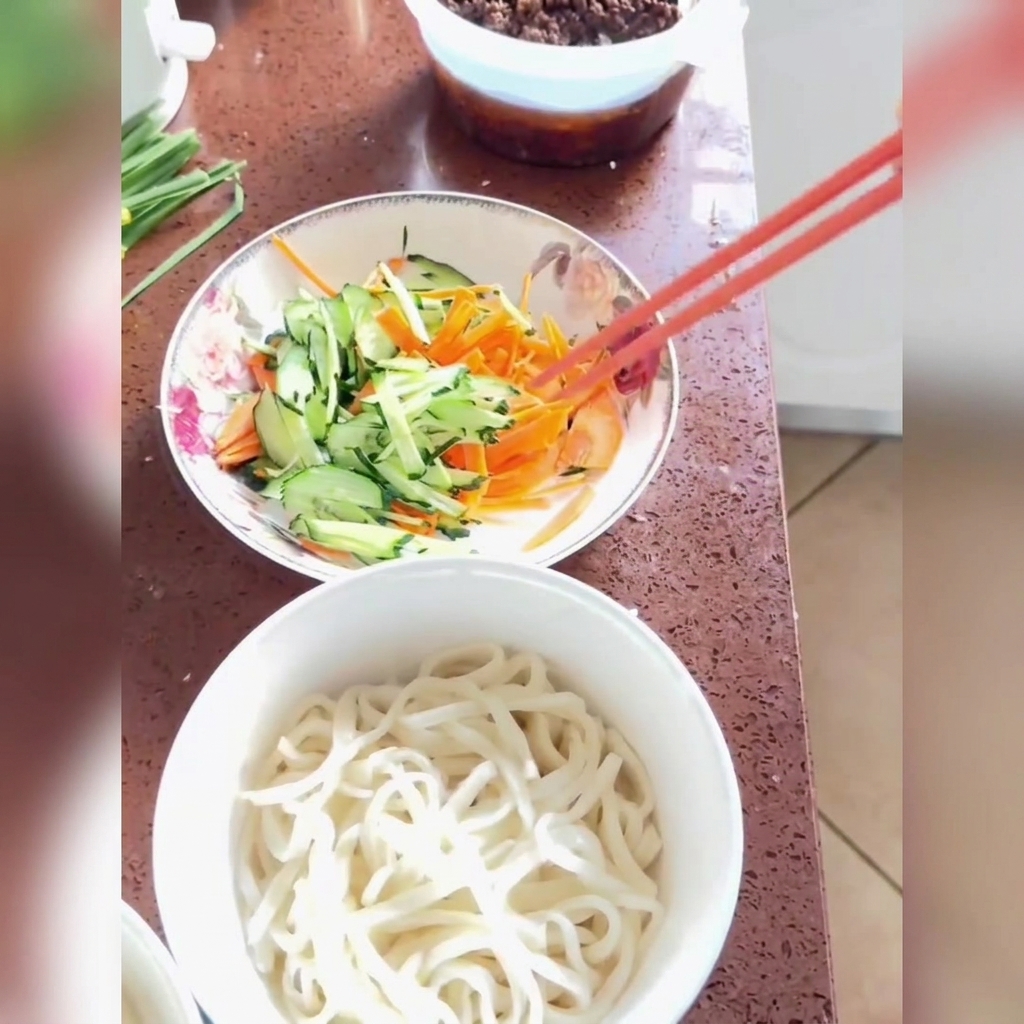 Salsa fritta versata e gocciolata sopra noodles appena cotti guarniti con cetrioli e carote tagliati a julienne in una ciotola bianca.