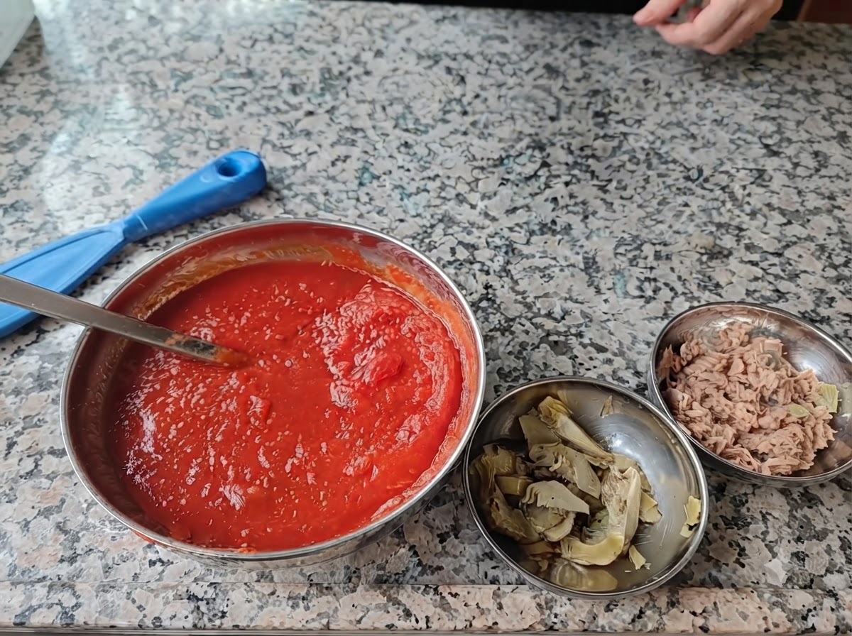 Quattro ciotole di acciaio piene di ingredienti per pizza disposte su una superficie di marmo.