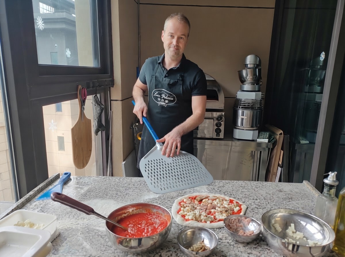 Un pizzaiolo che tiene una grande pala per pizza in metallo forato posizionata proprio accanto a una pizza cruda completamente assemblata.