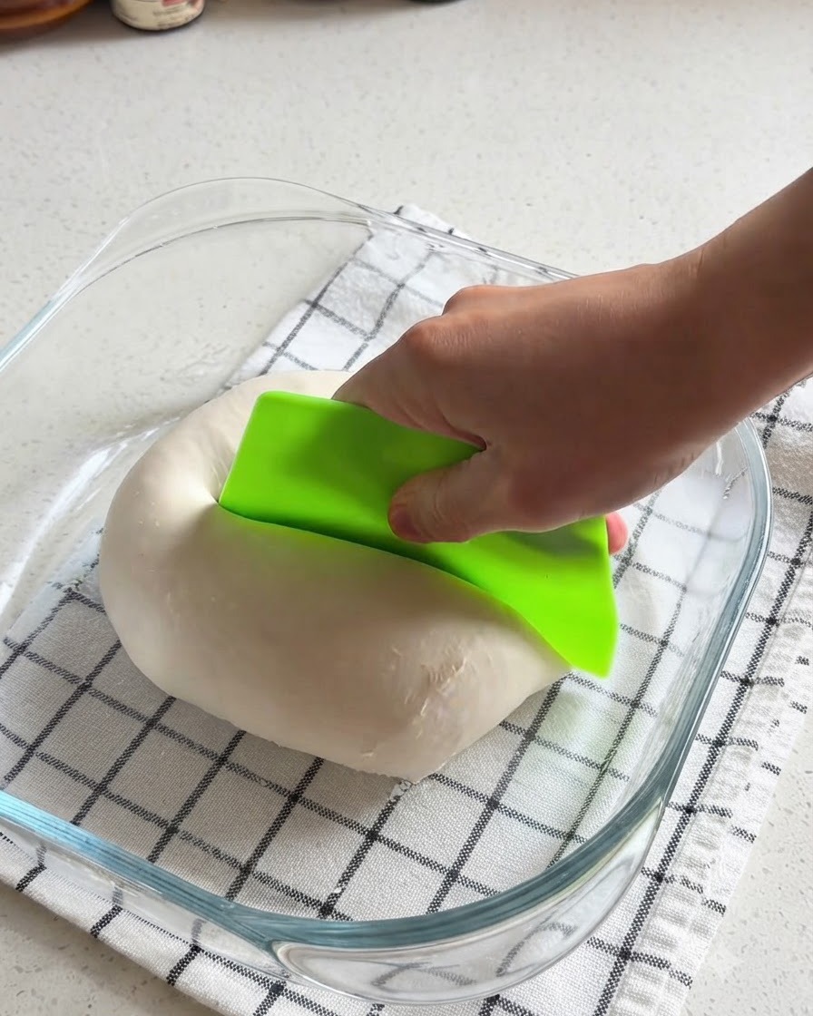 Una mano che utilizza un tarocco verde brillante per tagliare a metà una palla di impasto per pizza liscia allinterno di un recipiente di vetro.