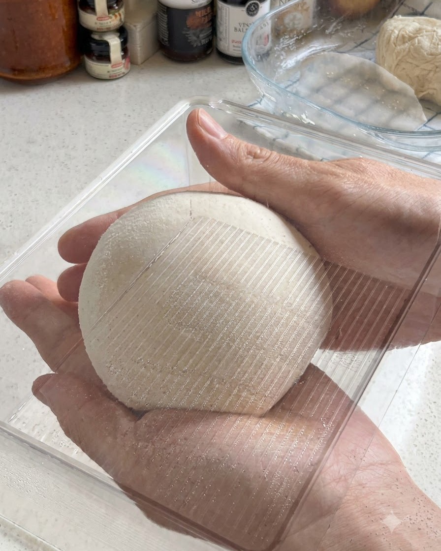 Due mani che tengono una palla di impasto per pizza perfettamente arrotondata mentre viene inserita in un contenitore di plastica trasparente per la lievitazione.