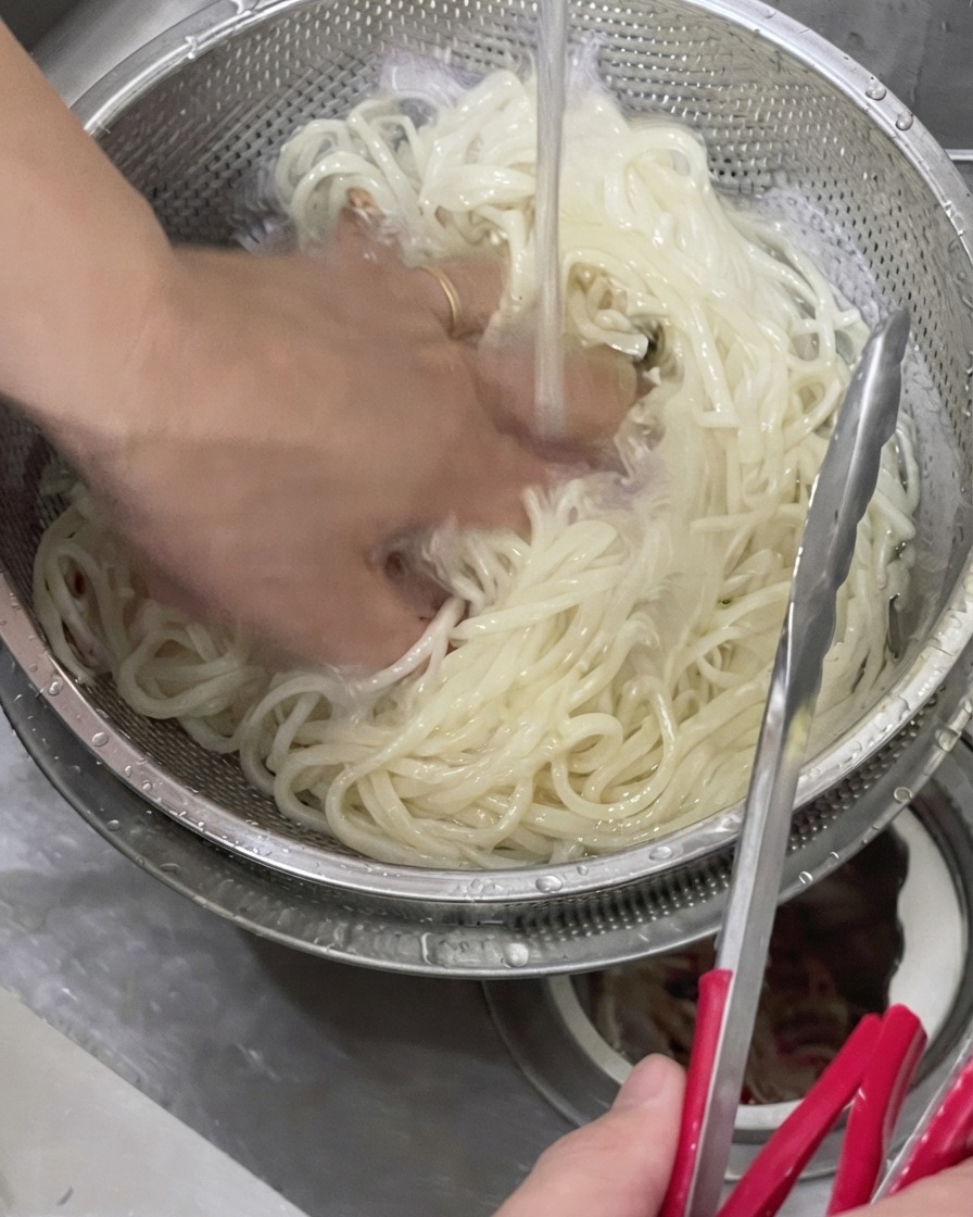 Una mano mescola gli udon cotti in un colino di metallo sotto lacqua fredda corrente.