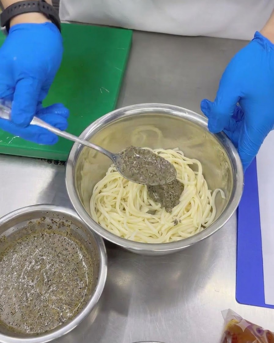 Mani con guanti usano un cucchiaio per aggiungere la densa salsa al tartufo nero agli udon freddi in una ciotola di metallo.
