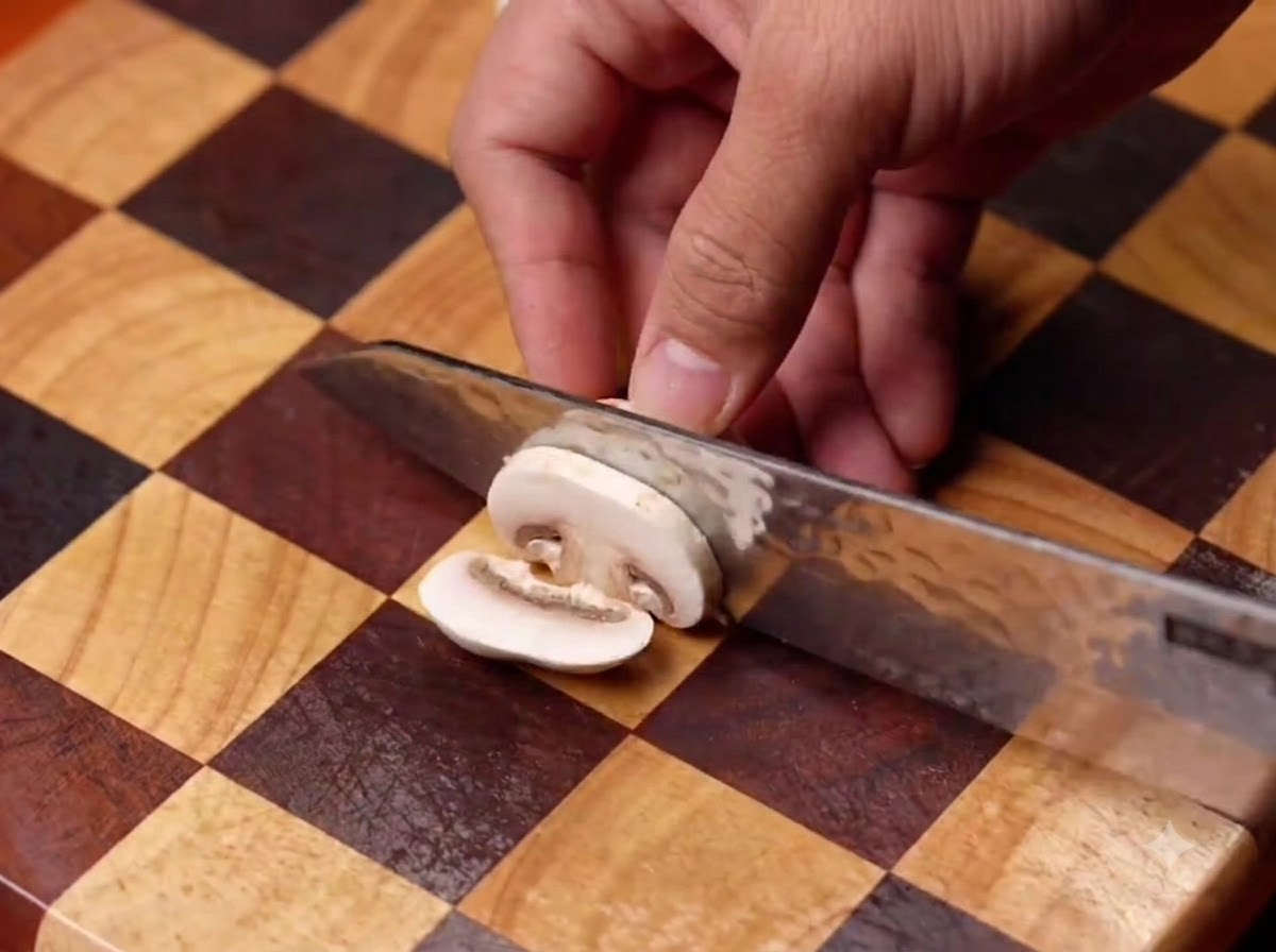 Funghi champignon affettati con un coltello su un tagliere di legno a quadretti, poi aggiunti in una padella nera.