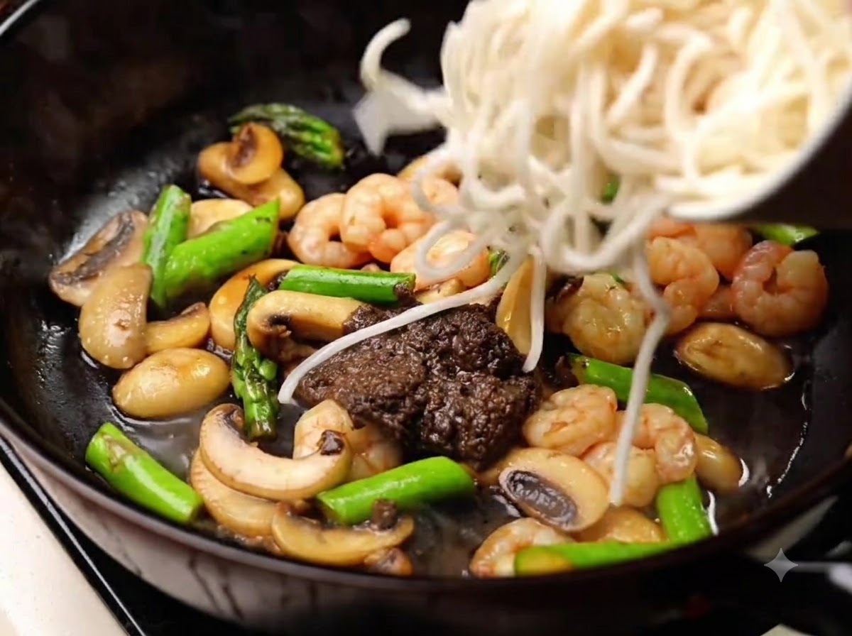 Noodles cotti aggiunti nella padella per essere mescolati con la salsa, i funghi, i gamberi e gli asparagi.