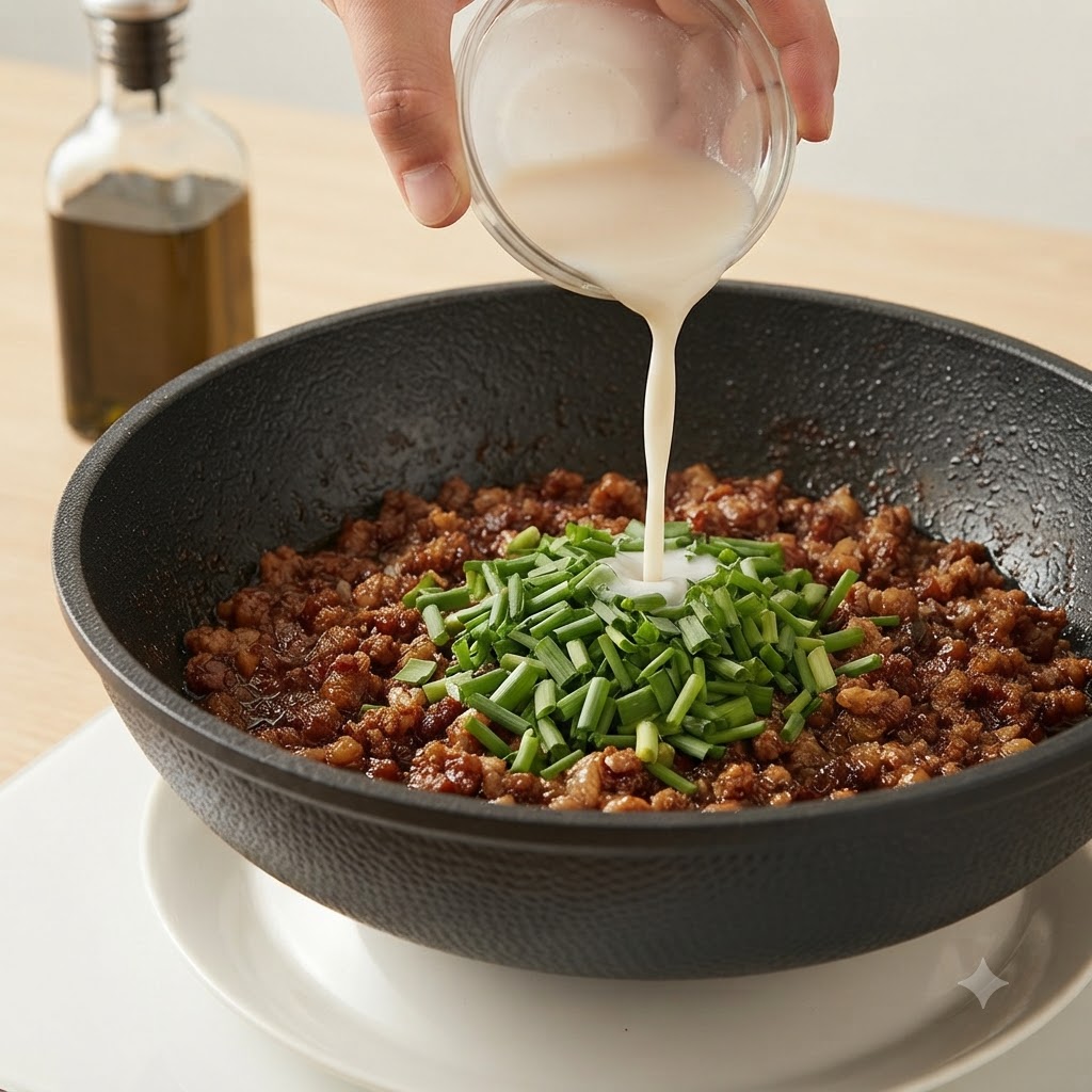 Una mano che versa una pastella di amido bianca da una ciotolina di vetro in una padella di carne macinata ed erba cipollina.