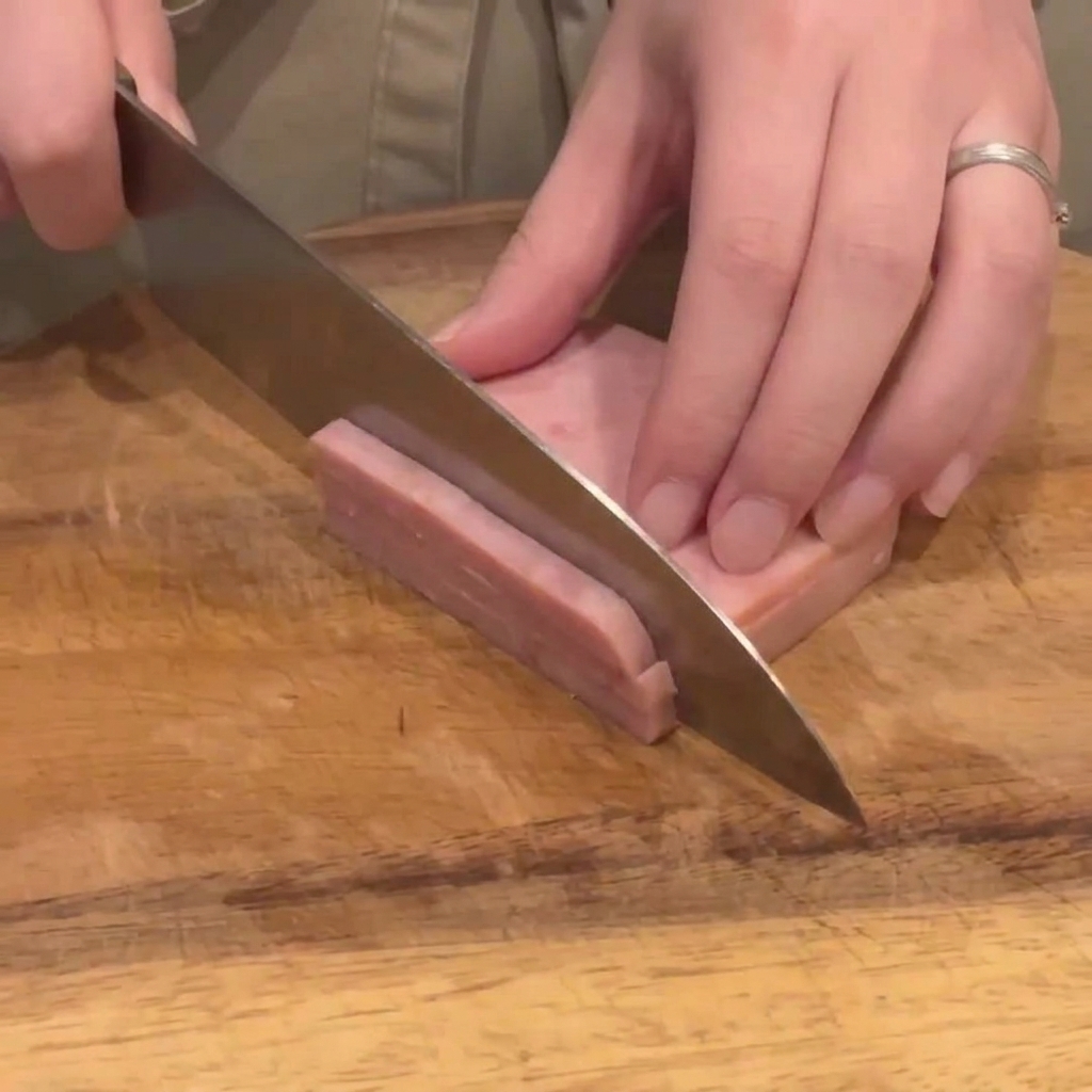 Un coltello da cucina affetta un blocco di prosciutto rosa in strisce sottili e uniformi su un tagliere di legno.
