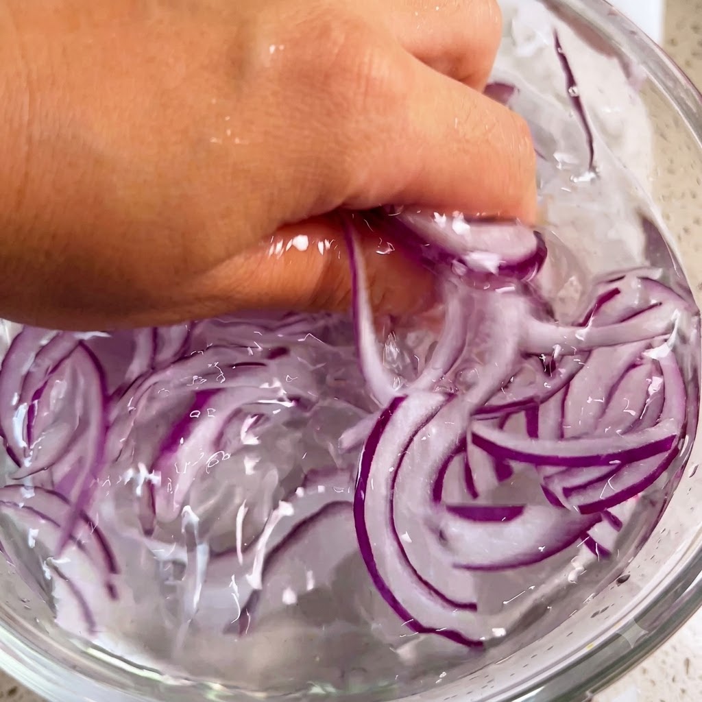 Una mano che lava delicatamente delle listarelle di cipolla viola allinterno di una ciotola piena di acqua ghiacciata.