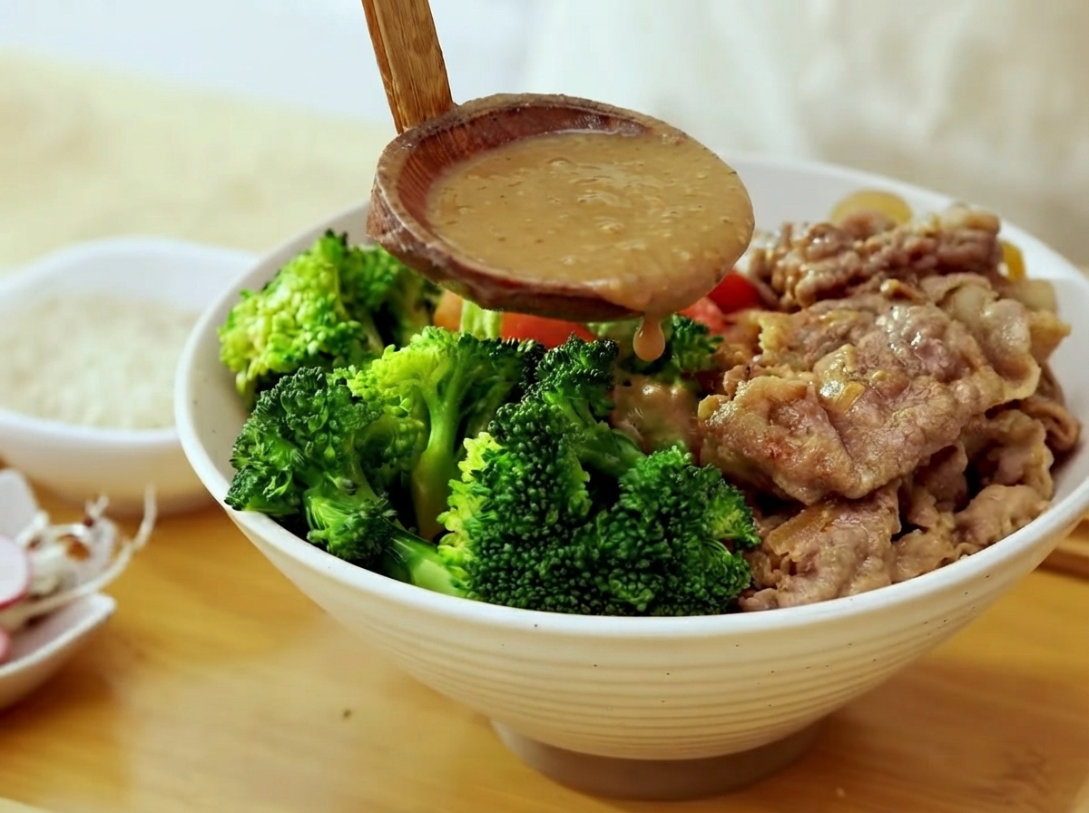 Un mestolo di legno che versa salsa densa al miso su una ciotola con broccoli, manzo cotto e riso.