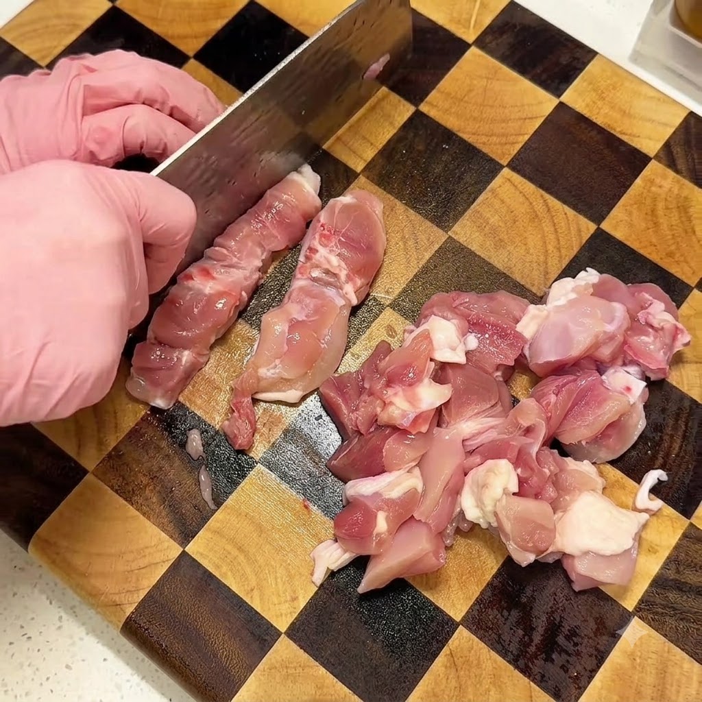 Mani con guanti rosa che usano una mannaia per tagliare cosce di pollo crude a dadini su un tagliere di legno a quadretti.