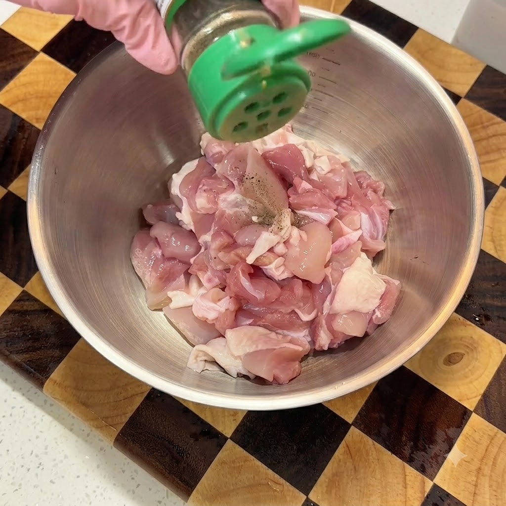 Spolverata di pepe nero in polvere da un contenitore verde sul pollo crudo a dadini in una ciotola di metallo.