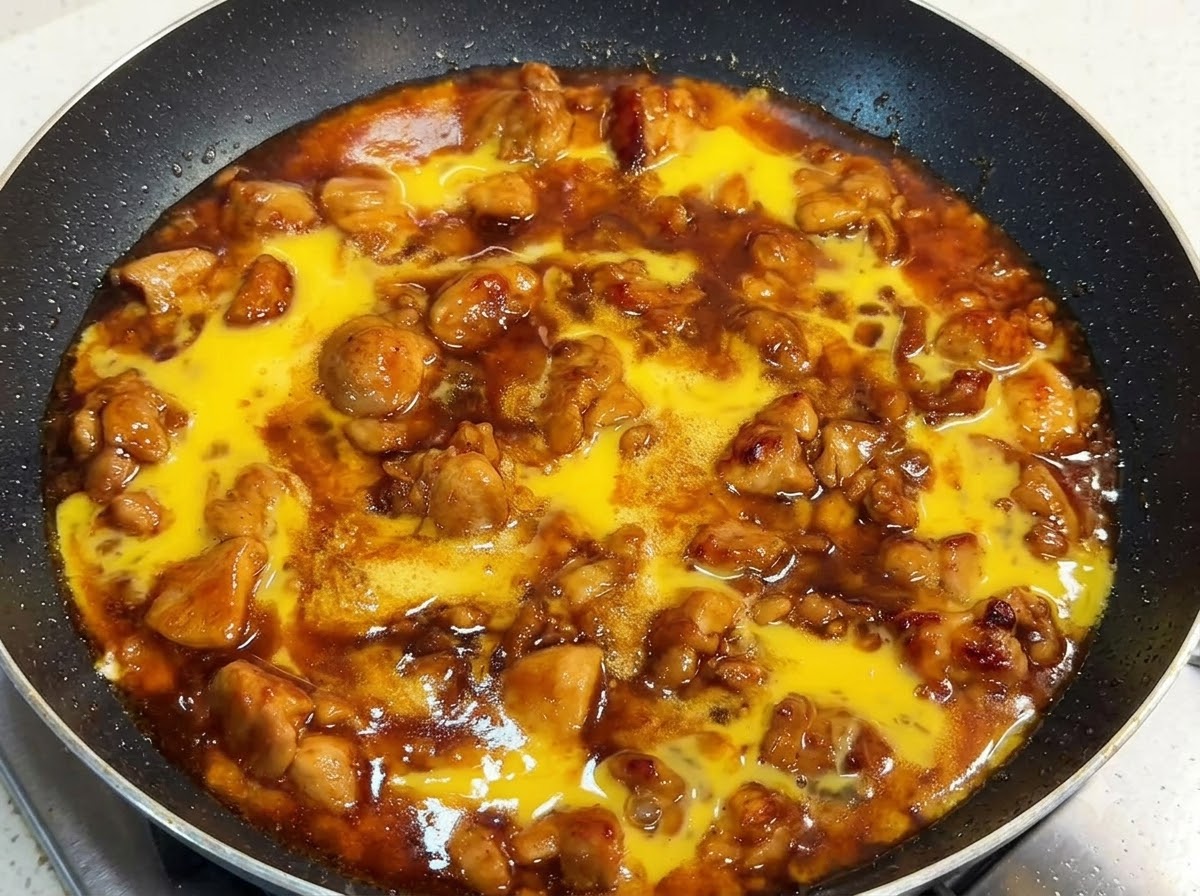 Una padella piena di pezzi di pollo e salsa ricca sormontata da liquido di uovo sbattuto crudo, coperta con un coperchio.
