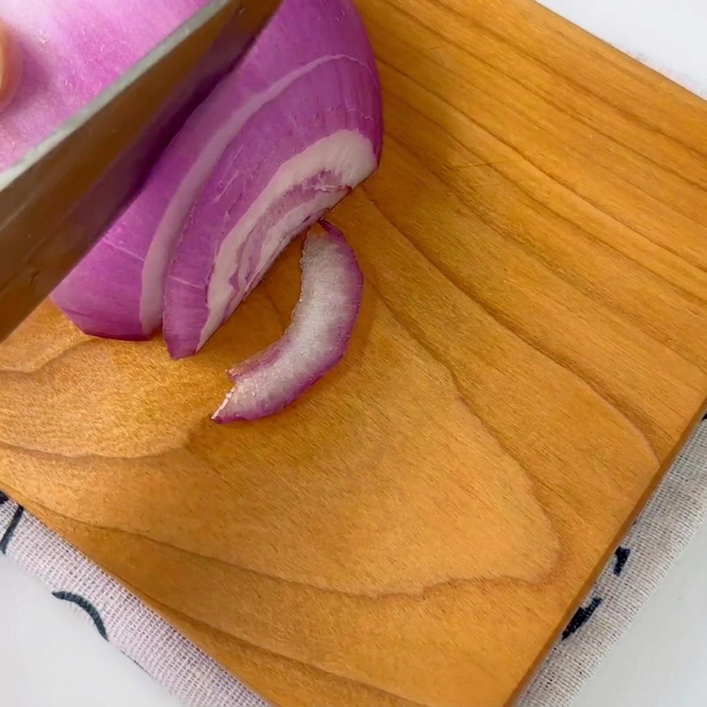 Una cipolla rossa che viene affettata con cura in strisce sottili su un tagliere di legno con un coltello.