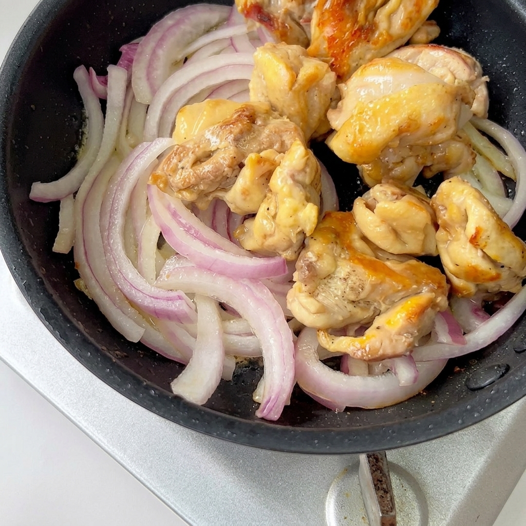 Pezzi di pollo cotto e cipolle rosse affettate che cuociono a fuoco lento in padella con la salsa.