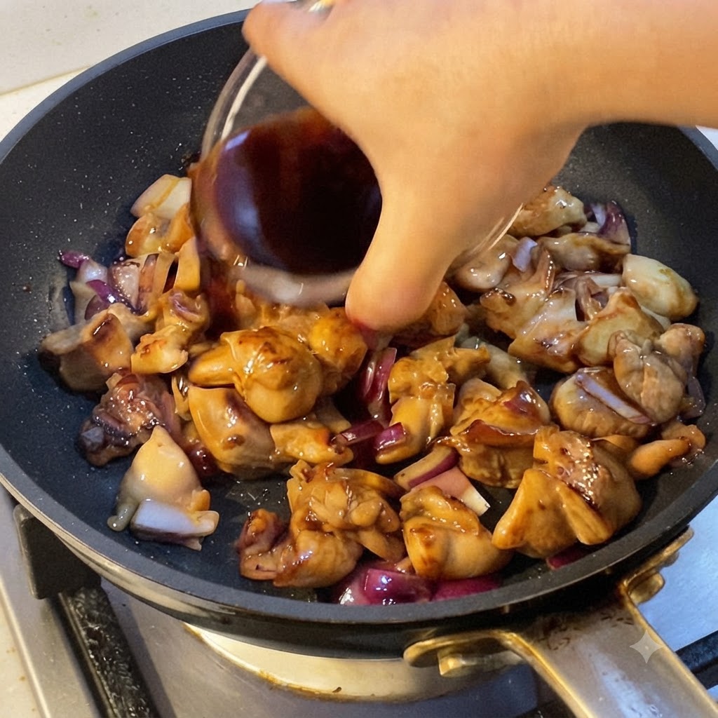 Una mano che versa la salsa di cottura scura in una padella piena di pezzi di pollo cotto e pezzi di cipolla rossa.