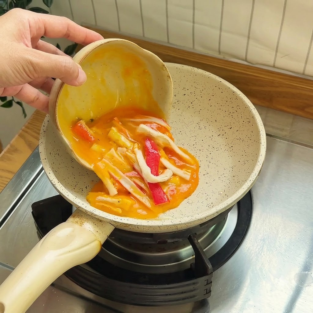 Uova sbattute mescolate con pezzi di surimi rossi e bianchi versate in una piccola padella antiaderente.