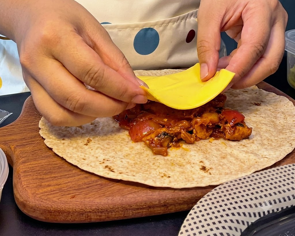 Mani che posizionano una fetta quadrata di formaggio giallo sopra un mucchio di ripieno caldo di pollo e kimchi su una piadina.