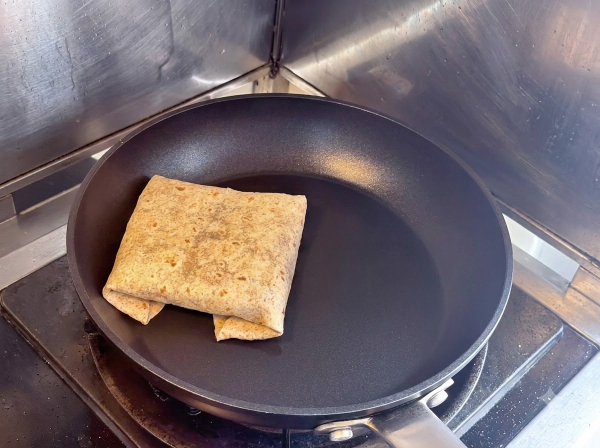 Un wrap a piadina quadrata piegato che tosta con la cucitura rivolta verso il basso in una padella antiaderente nera asciutta.