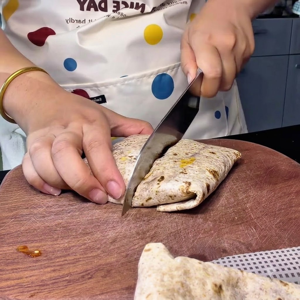 Una persona che usa un grosso coltello da cucina per tagliare un wrap integrale disposto diagonalmente su un tagliere di legno.