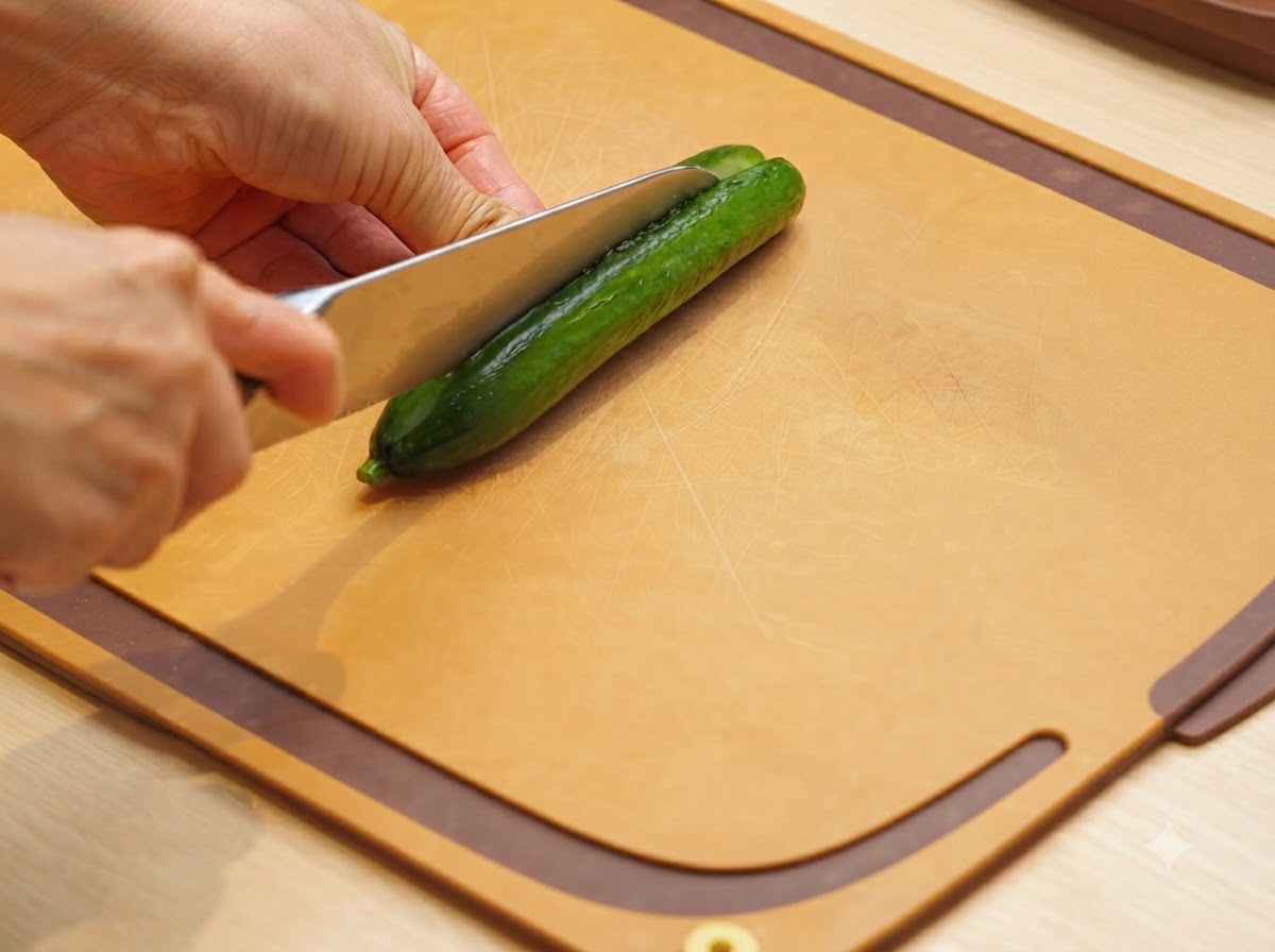 Un coltello da chef affilato che taglia un cetriolo per il lungo su un tagliere marrone.