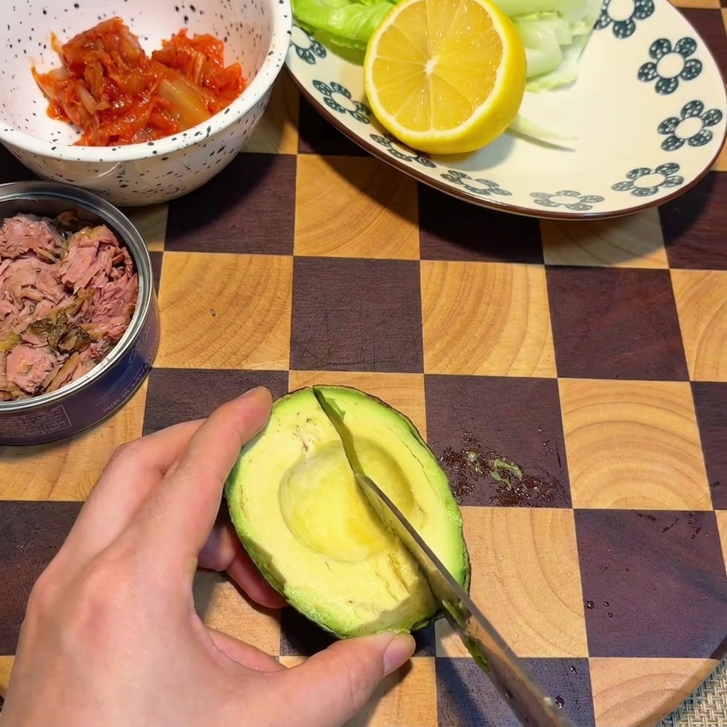 Una mano che usa un coltello per tagliare cubetti a griglia in una metà di avocado appoggiata su un tagliere di legno a quadretti.