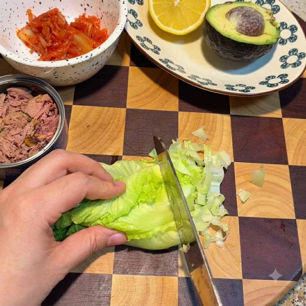 Un coltello che taglia foglie di lattuga verde fresca su un tagliere a quadretti accanto a una ciotola di kimchi e una scatola di tonno.