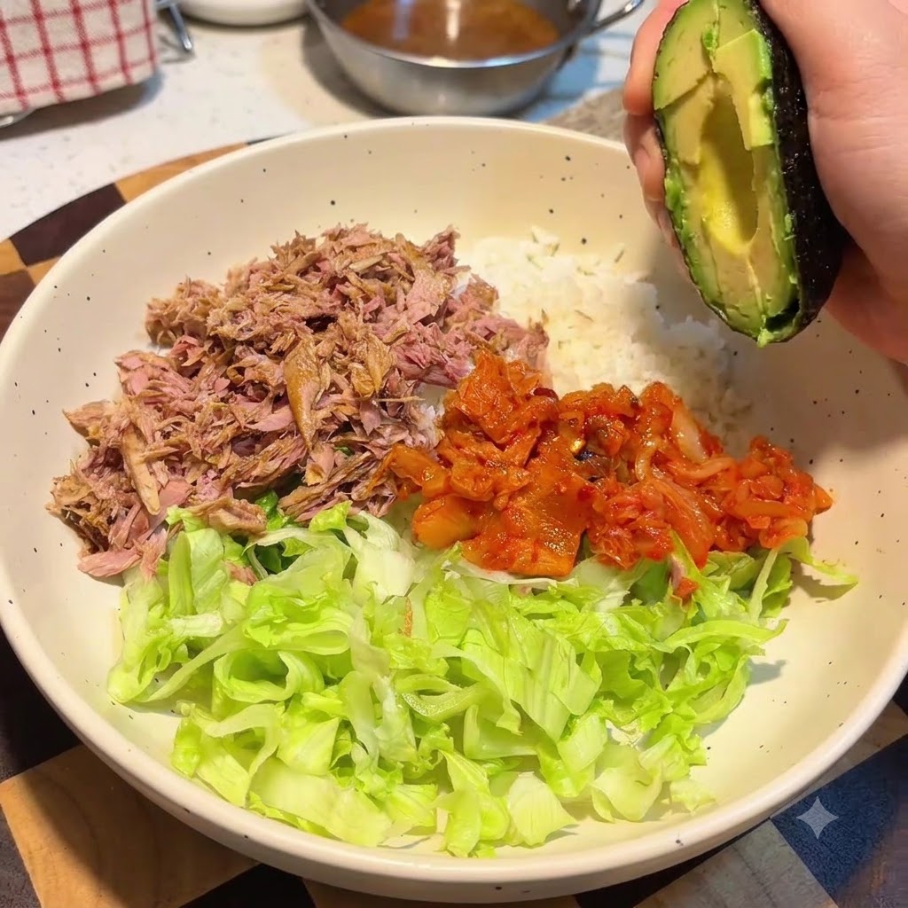 Una mano che tiene una metà di avocado sopra una ciotola grande contenente riso, lattuga, tonno e kimchi.