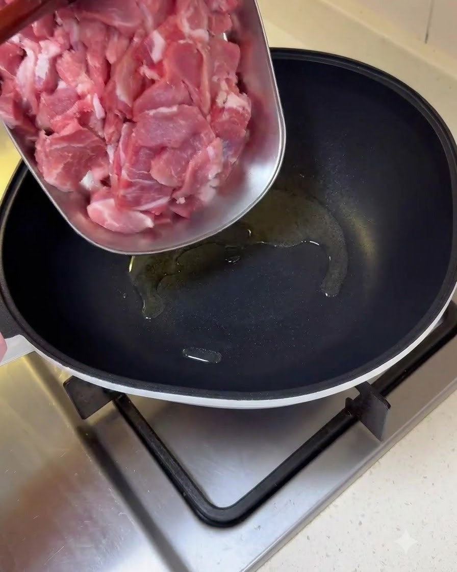 Pezzi di maiale crudo affettati trasferiti da un vassoio a una padella oliata.