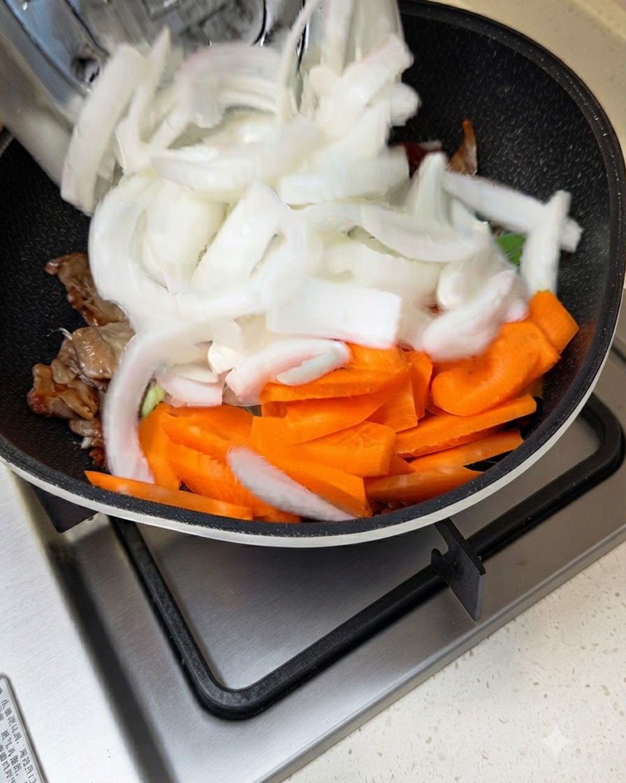Cipolle bianche e carote affettate aggiunte in una padella con carne e salsa in fase di cottura.