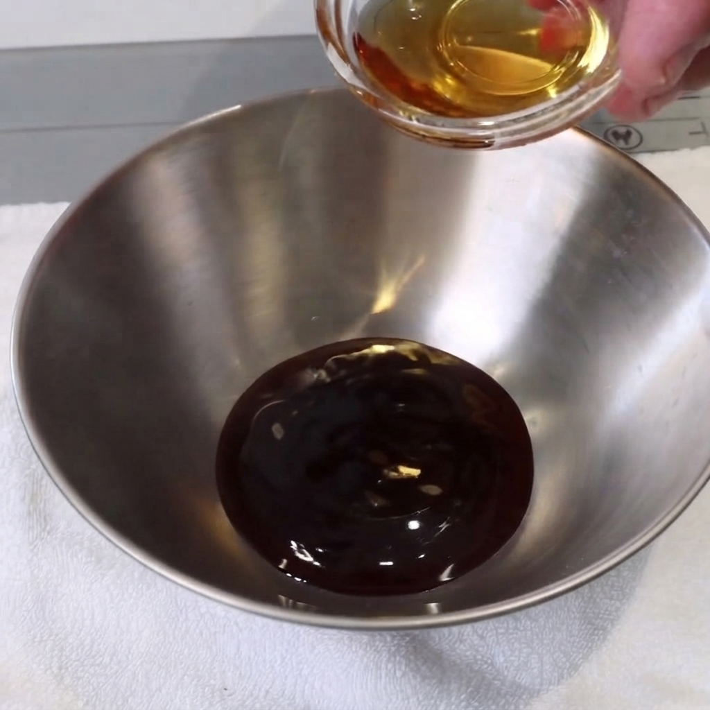 Una piccola ciotola di metallo con ingredienti liquidi mescolati con un cucchiaio su un telo bianco.