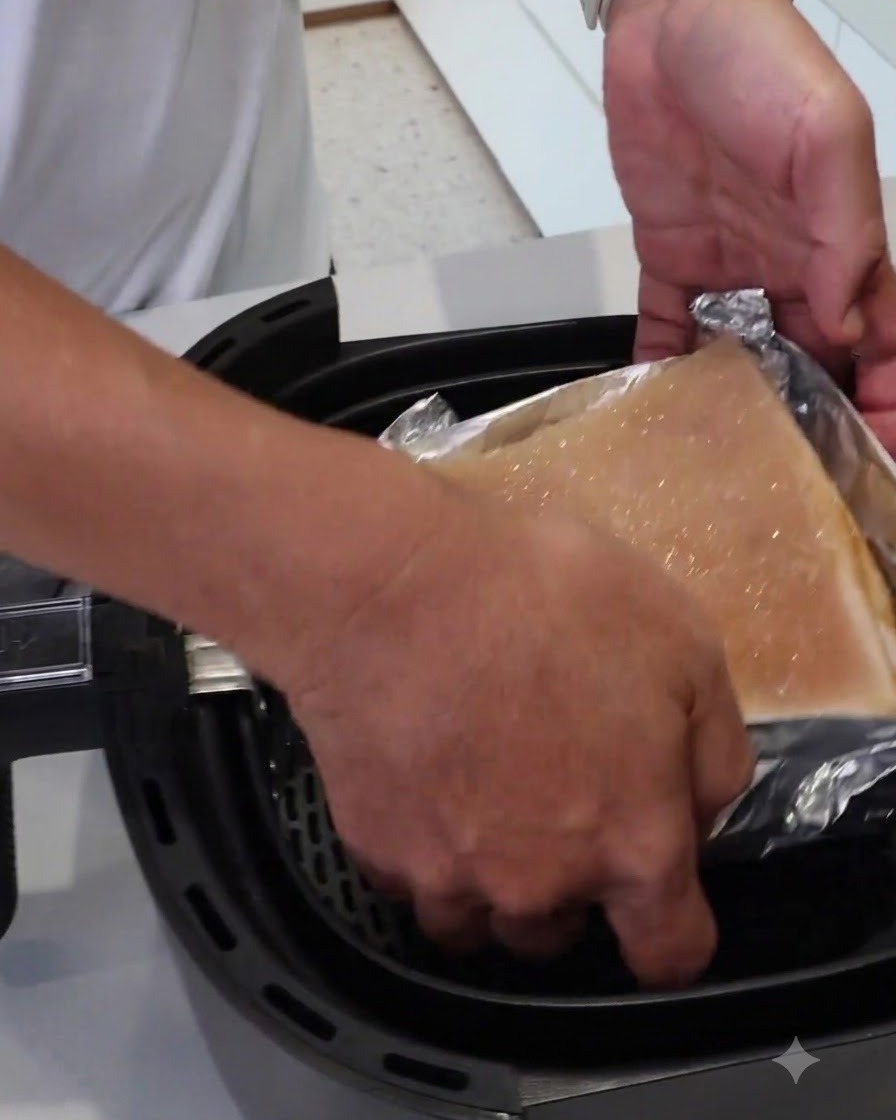 Una coppia di mani che cala un blocco di carne avvolto in alluminio in un cestello per friggitrice ad aria.
