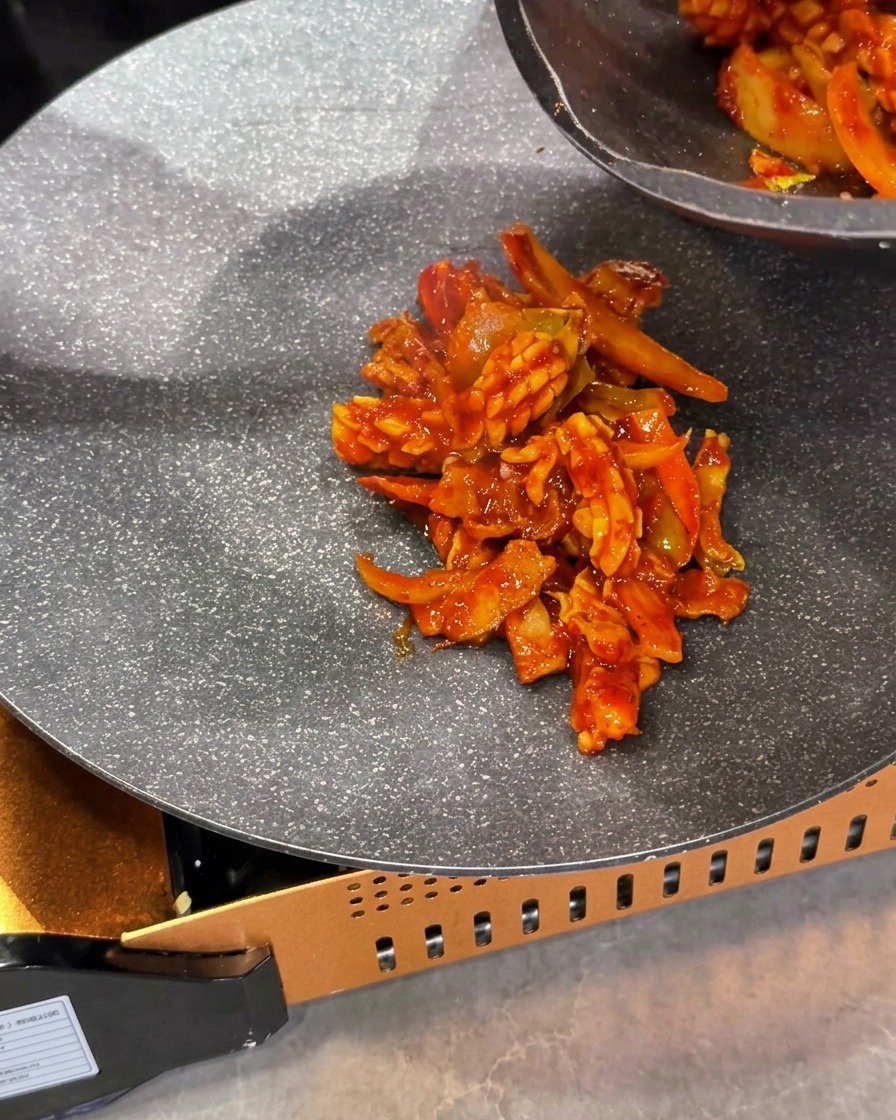 Il composto cotto e rosso di totano e maiale saltato posizionato al centro di una padella su fornello a gas portatile.