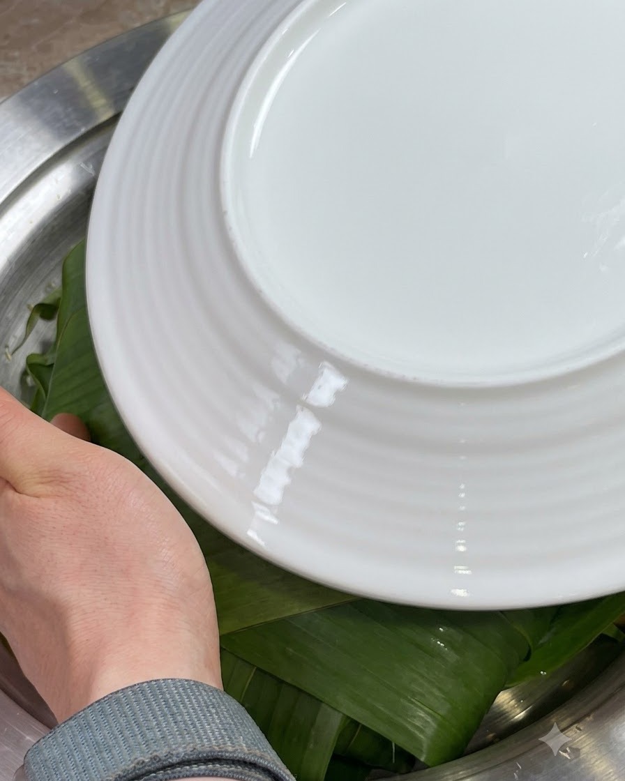 Mani che piegano i bordi delle foglie di Zong verdi verso linterno sopra gli ingredienti, coperti con un piatto bianco.