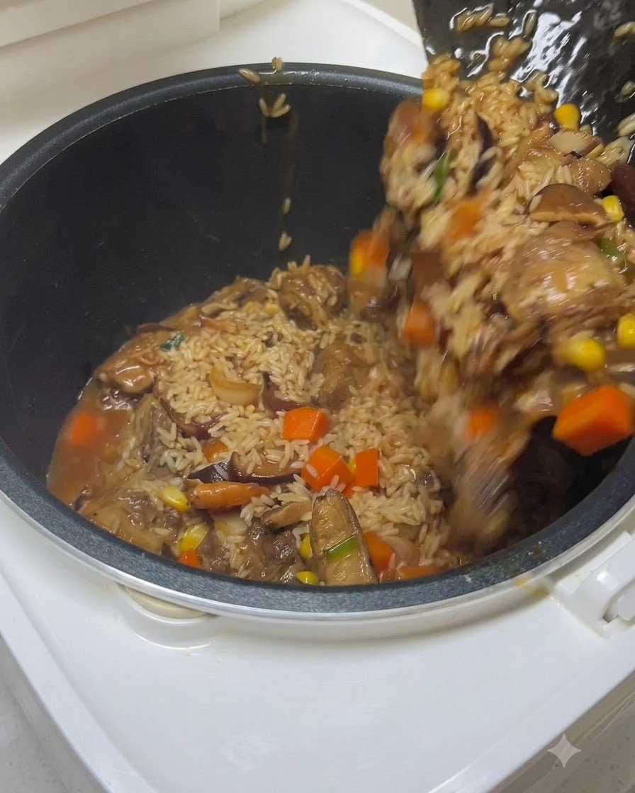 Una spatola che trasferisce riso condito, costine e verdure in una pentola del cuociriso mentre viene versata dellacqua.