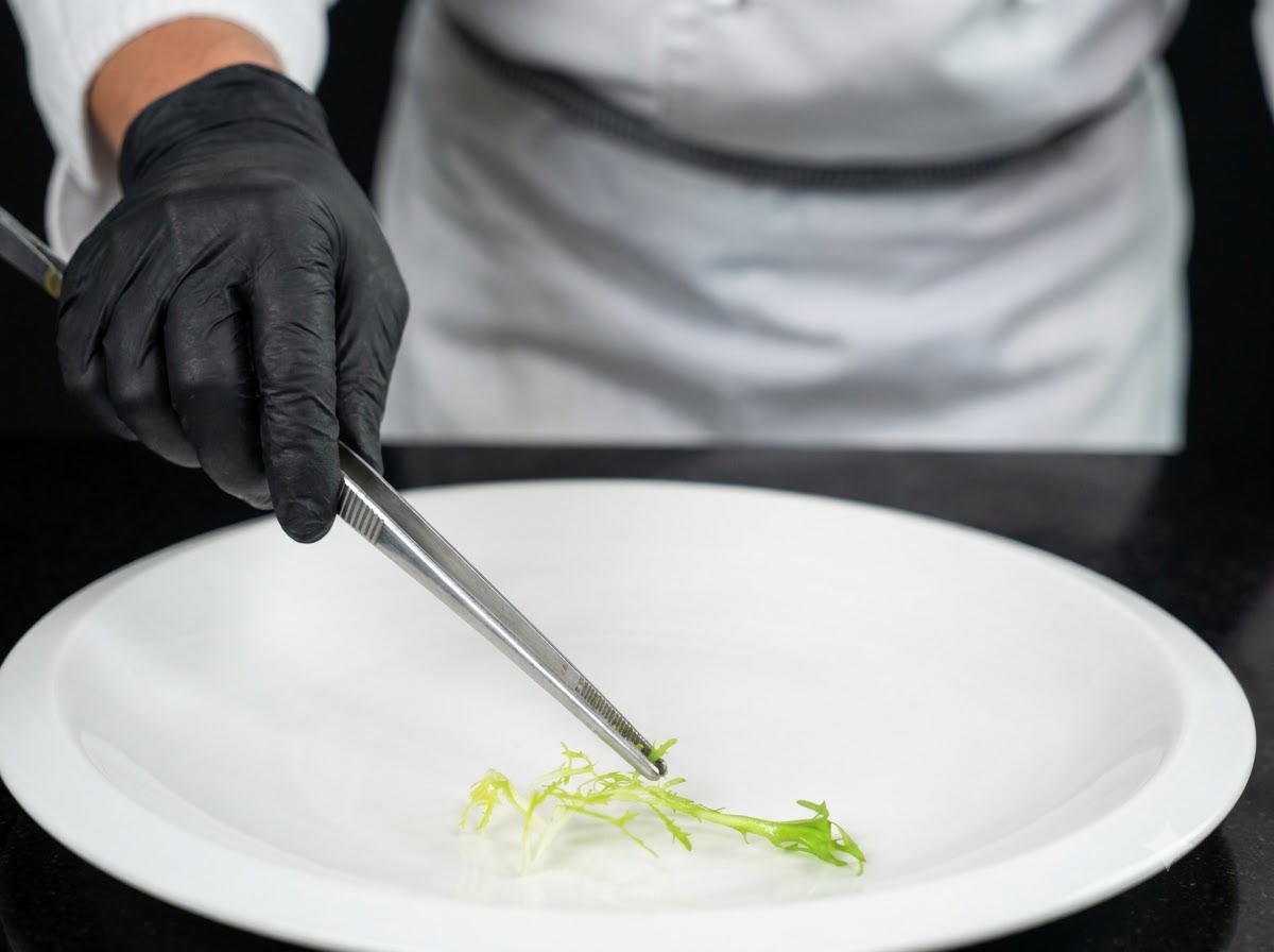 Uno chef che indossa guanti neri usa le pinze per posizionare un contorno di verdure verdi fresche al centro di un grande piatto bianco.