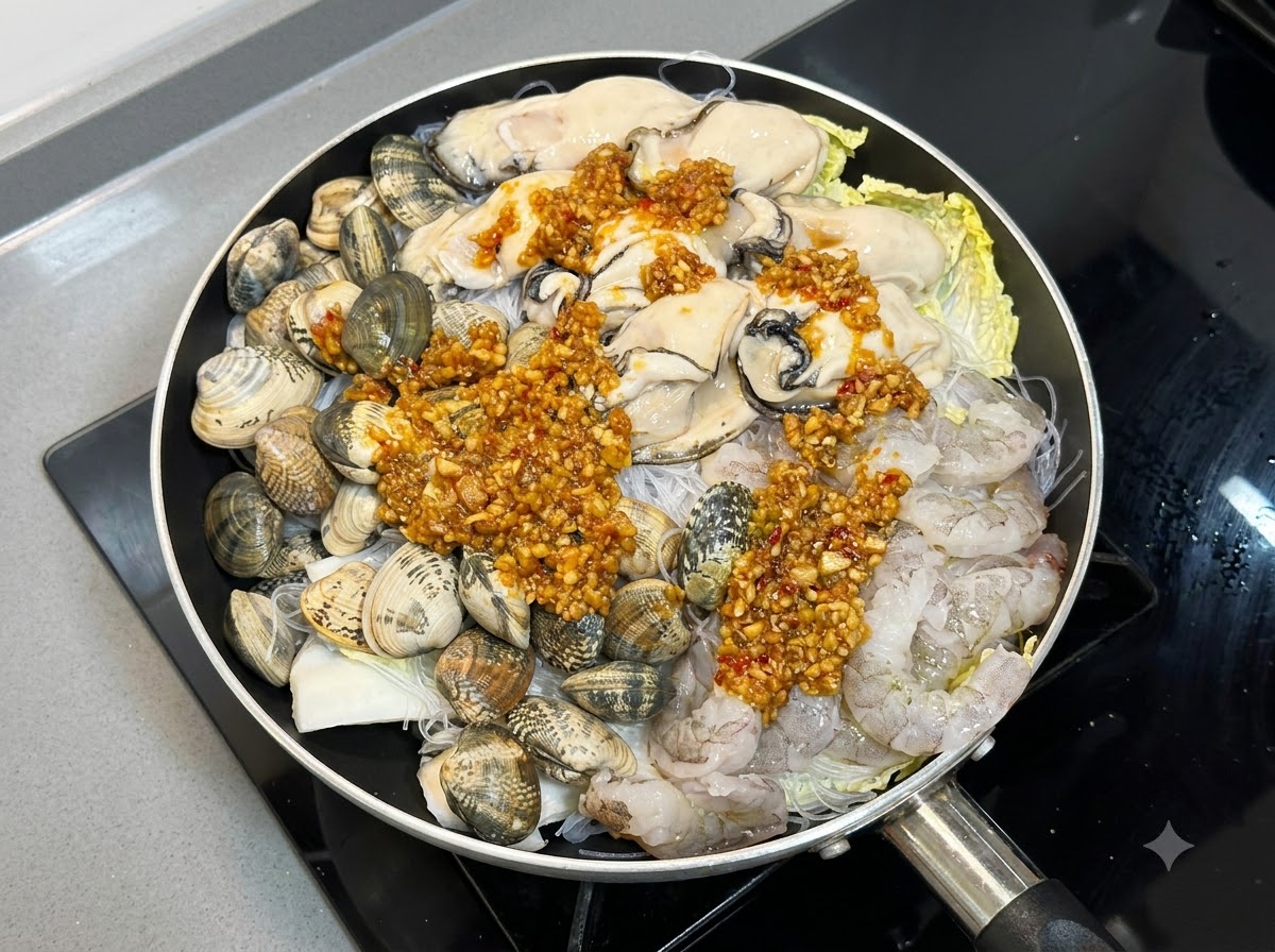 Salsa cotta di aglio e peperoncino generosamente cucchiaiata sugli strati crudi di frutti di mare e noodles in una padella.