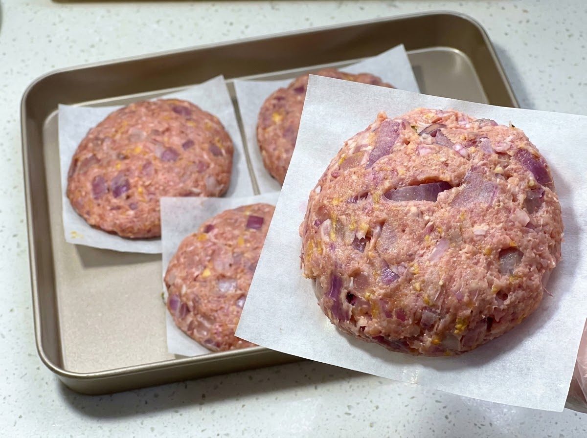 Un hamburger crudo appena formato su carta da forno, con altri hamburger disposti su un vassoio dietro.