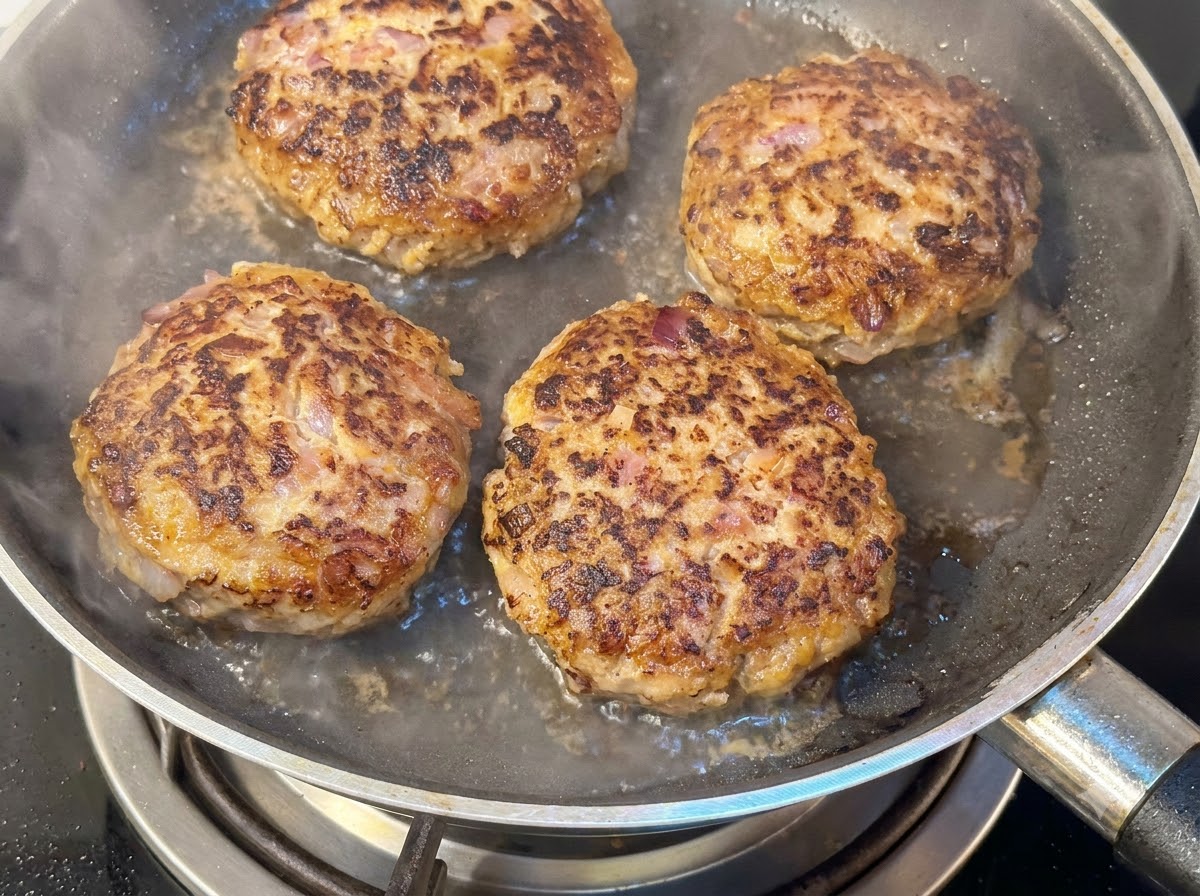 Quattro spessi hamburger che friggono in padella con acqua e vapore che sale sotto un coperchio.